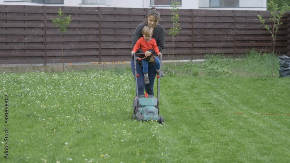 Young mother holding baby child working in garden with lawn mower Young mother holding baby child working in garden with lawn mower