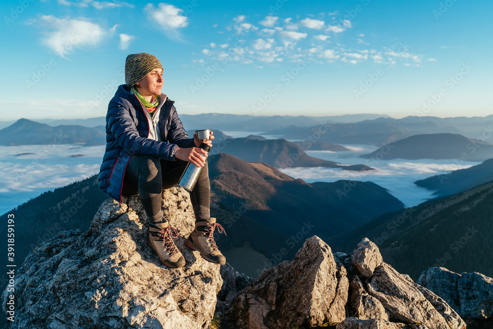 Young hiker woman sitting on the mountain summit cliff, drinking tea from a thermos flask and enjoying mountains valley covered with clouds view. Successful summit climbing concept image. - obrazy, fototapety, plakaty 