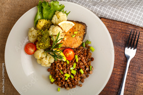 Baked fish with broccoli and lentils in a white plate with rosemary and lemon on a napkin and a round stand next to a fork.