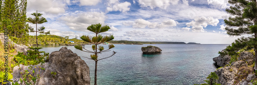 Amazing panoramic shot of Island of Mare in New Caledonia. Lone massive cliff in the water next to the island. Trees and rocks in the foreground. Blue cloudy sky as a background
