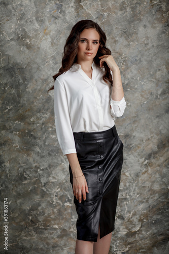 A beautiful girl with long hair in a black leather skirt and white blouse. Business style clothing. Business woman in a formal attire stands in the light room.