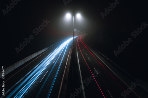 Horizontal photo of cars light trails on a curved highway at night with street lamps over the road. Motion blur image - city road with traffic headlight motion (top view) in foggy and misty weather.