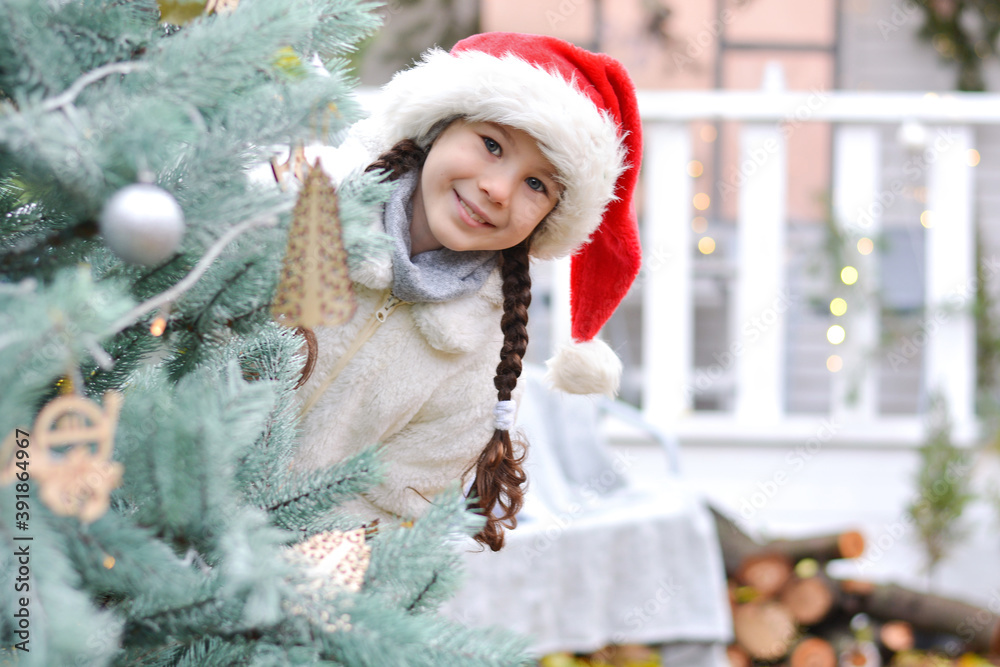 A happy little girl in a white fur coat and santa hat peeks out from behind a Christmas tree against the backdrop of a New Year's house. Holidays and people concept A happy little girl in a white fur coat and santa hat peeks out from behind a Christmas tree against the backdrop of a New Year's house. Holidays and people concept