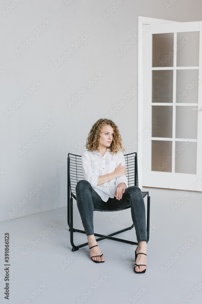 A beautiful blonde girl in a white shirt and jeans is sitting on a chair on a gray background in the studio. A beautiful blonde girl in a white shirt and jeans is sitting on a chair on a gray background in the studio.