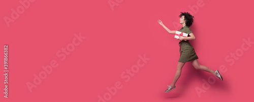 curly haired woman is running after somebody holding a present and gesturing with hands the wait sign on red wall with free space