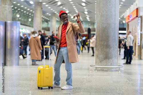 Afro-American traveler millennial man with yellow suitcase stands in airport terminal after arrival, holding mobile phone and paper cup of coffee, raising hand, greeting someone with hand, say hello