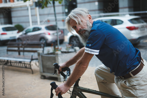 Elderly grey haired bearded man looking to the camera, riding bicycle on city streets