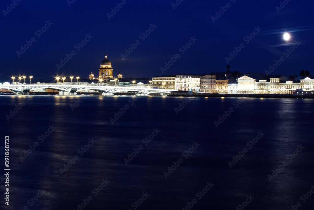 A dark night river against the backdrop of a panorama of the city brightly lit by lights, shooting at night.