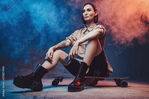 Sportive and stylish woman with tattoo and short haircut sits on skateboard holding her smartphone in smokey background.