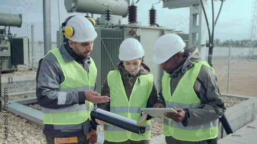 Multinational construction engineers in helmets hold tablet and folder discussing project confidently at power station close view