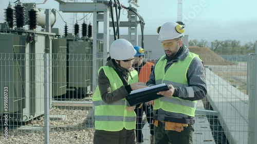 Power engineers man and lady taskmaster in protective helmets hold black folder and discuss project against electric substation
