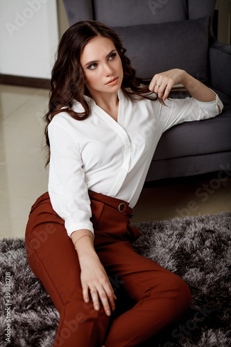 A young beautiful girl with a beautiful hairstyle and makeup in a white blouse and red trousers sitting on the floor, leaning on a chair. Portrait of a model in office style clothes.