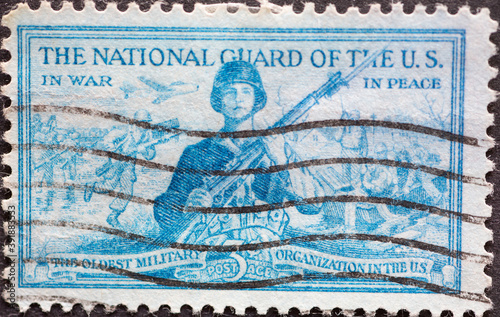  a postage stamp printed in the US showing a National Guardsman ready for action. The two background images picture a guardsman in a war scene and another protecting life and property.