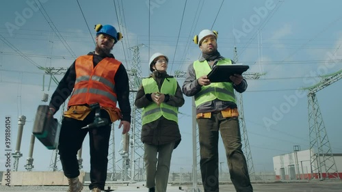 Construction engineers and woman brigadier in helmets walk along electric power substation and discuss project low angle shot