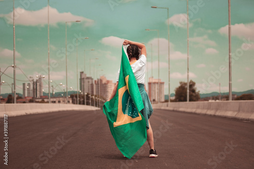 Mulher em cima de uma ponte, com a cidade ao fundo, trajando blusa branca e camisa na cintura, segurando a bandeira do Brasil e sorrindo photo