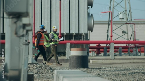 Skilled bearded power electric station workers and lady engineer in protective helmets walk discussing project on sunny autumn day