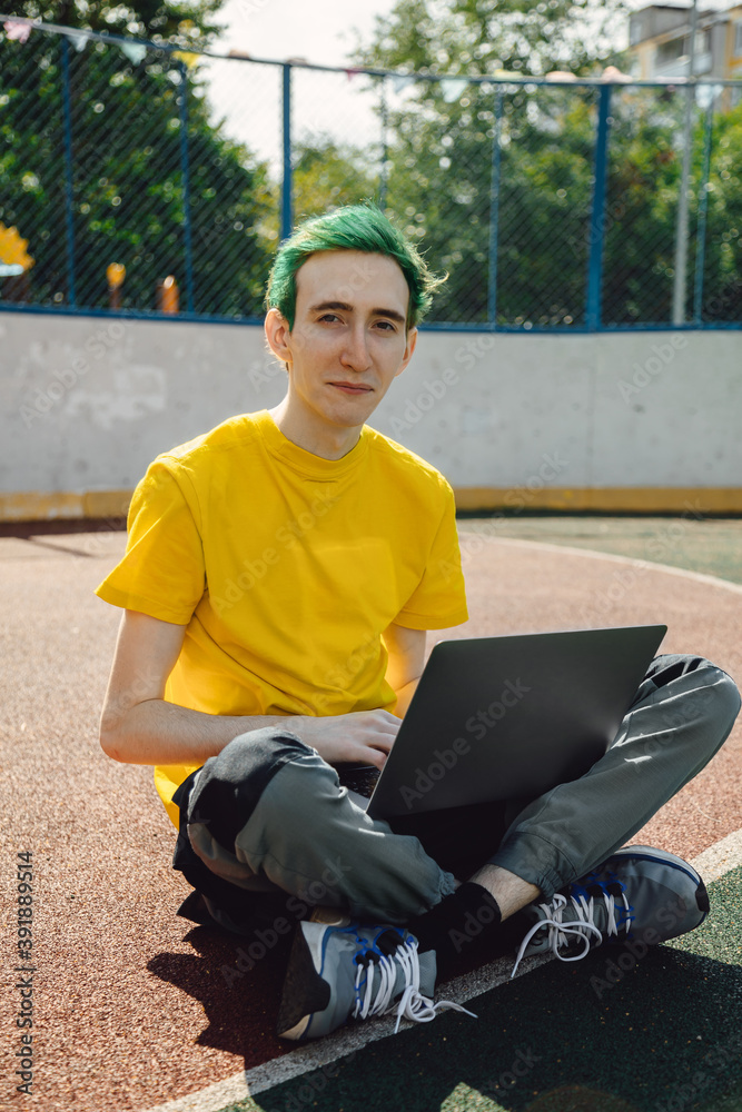 Fototapeta premium Freelancer, it professional in free space, remote working at online platform for programmers. Smiling young male freelancer with green hairstyle working on laptop in outdoor street.