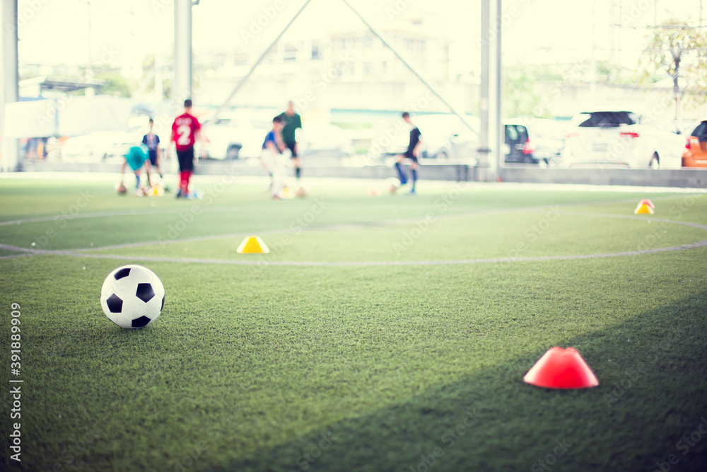 Obraz premium Soccer ball on green artificial turf with blurry kid soccer team training. Blurry kid soccer player jogging between marker cones and control ball with soccer equipment in football academy.