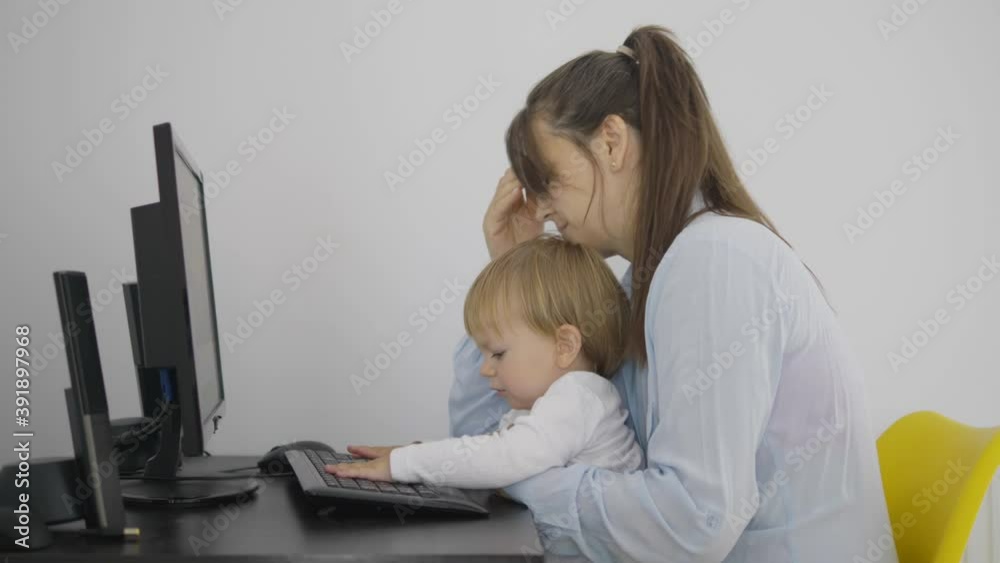 Mother holding a baby child working at computer, tired woman, boy taping Mother holding a baby child working at computer, tired woman, boy taping
