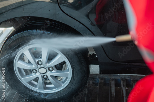 A car cleaner wearing a red uniform and a standing hat sprays water using a hose onto the tires in the car wash