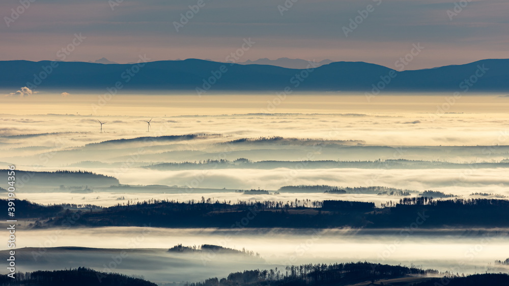 Sun light rays during sunshine peaking through morning mist on a wavy landscape with wind turbines peaking out.