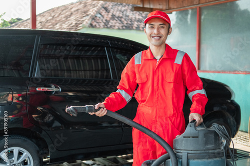 male car cleaner wearing red uniform standing carrying vacuum cleaner in car salon