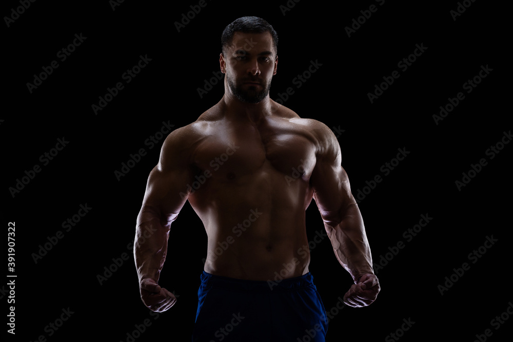 Portrait of a bodybuilder standing isolated on black background in a shadow with clenched fists to show off his muscles
