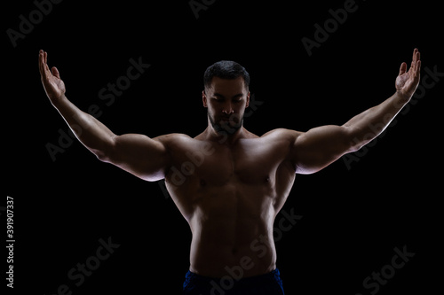 Portrait of a bodybuilder standing isolated on black background in a shadow with raised hands to show off his muscles