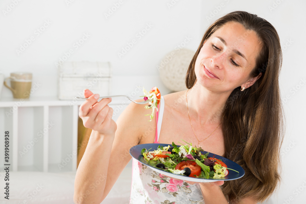 Positive pretty woman holding fork and eating vegetable salad in bed at home