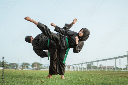 Asian male and female fighters wearing pencak silat uniforms face each other in an upward kick pose in an outdoor background photo