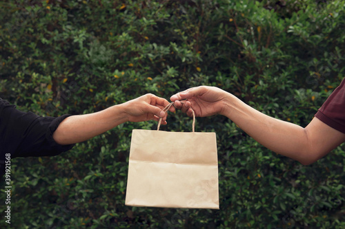 Woman’s hand taking empty craft brown recycled paper shopping bag from man with outdoor green leaves nature background. Delivery paper bag mockup for branding design concept photo