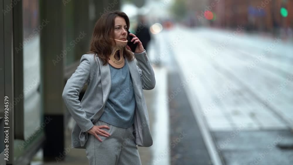 Portrait of a middle-aged woman, she has a corset around her neck, she is on the street in the city. wearing a gray jacket and pants. Holds the phone to his ear, standing by the road at the bus stop. Portrait of a middle-aged woman, she has a corset around her neck, she is on the street in the city. wearing a gray jacket and pants. Holds the phone to his ear, standing by the road at the bus stop.