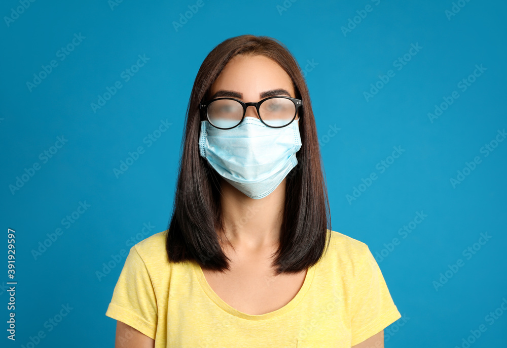 Young woman with foggy glasses caused by wearing disposable mask on blue background. Protective measure during coronavirus pandemic