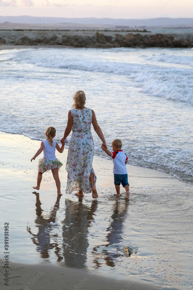 Happy family - young mother, daughter, baby son hold hands, run together with splashes by water pool along sea surf on white sand beach.