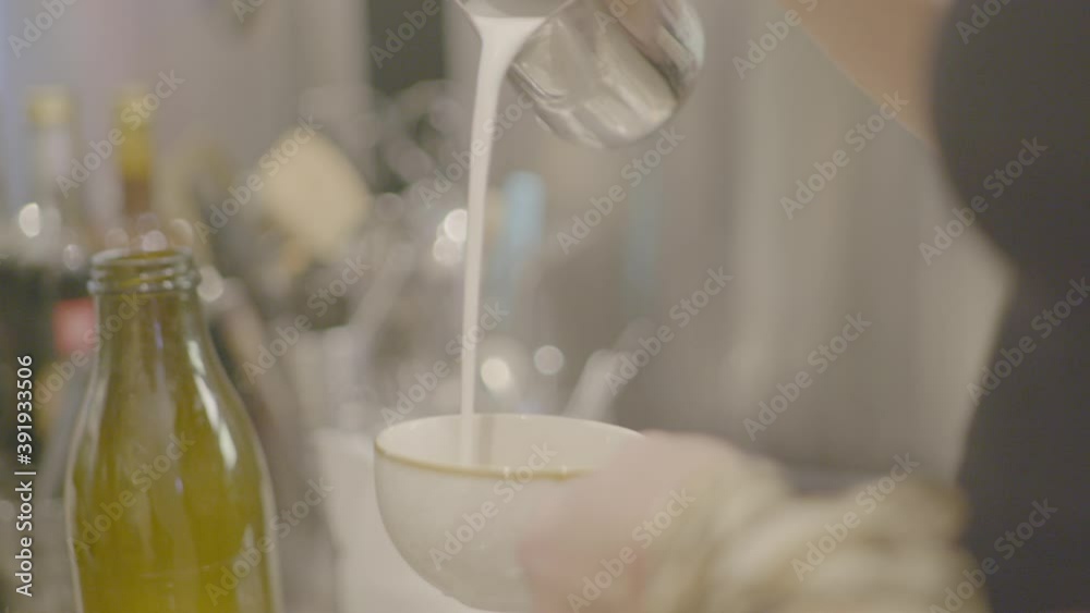 Barista slowly pouring a cup of milk into a cup of coffee with more cans and a coffee machine in the blurred background. Shot in slow motion.