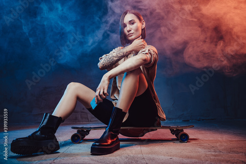 Brown haired beautiful girl with tattoo and smartphone poses in smokey background sitting on skateboard.