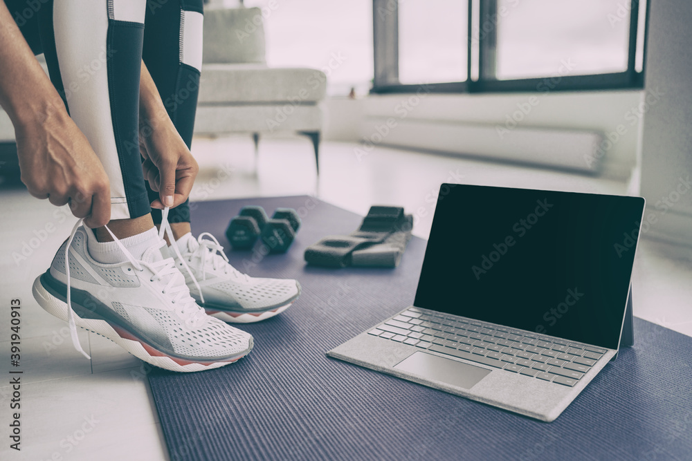 Fototapeta premium Home workout with laptop. Online fitness class . Woman getting ready for exercising tying her running shoes with technology device streaming on black screen.