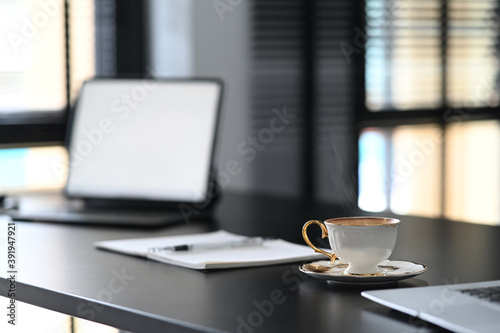 A white coffee cup is putting on contemporary workspace with empty screen computer laptop on blace wooden table.