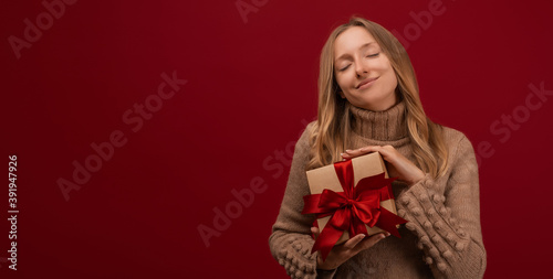 Charming blonde woman holding gift with red ribbon. Studio shot red background. Mock up copy space