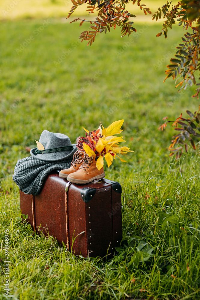 Autumn coziness, yellow and red foliage. Old boots, felt hat, knitted throw on the brown vintage suitcase in autumn forest. Shoes with foliage inside.