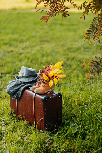 Obraz na płótnie Autumn coziness, yellow and red foliage. Old boots, felt hat, knitted throw on the brown vintage suitcase in autumn forest. Shoes with foliage inside. w salonie