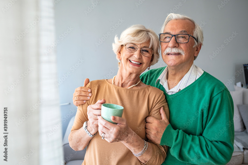 Happy senior man embracing woman in front of windows at home. Senior couple smiling and happy at home. Happy pensioner couple enjoying time at home Happy senior man embracing woman in front of windows at home. Senior couple smiling and happy at home. Happy pensioner couple enjoying time at home