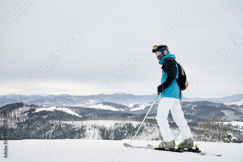 Full length man skier looking at beautiful snowy mountains. Young man wearing ski suit, backpack, protective helmet, gloves and winter ski pants. Concept of winter sport activities.