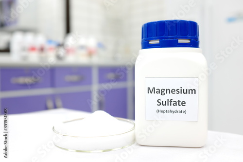 Selective focus of magnesium sulfate or epsom salt chemical compound bottle beside a petri dish with white crystalline powder in a chemistry laboratory background.  photo