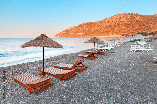 Fototapeta Naklejka Na Ścianę i Meble -  Sunbeds and sun umbrellas await vacationers on the shingle beach at Ovabuku beach on the Datca Peninsula in Turkey. The photo was taken in the early morning at sunrise