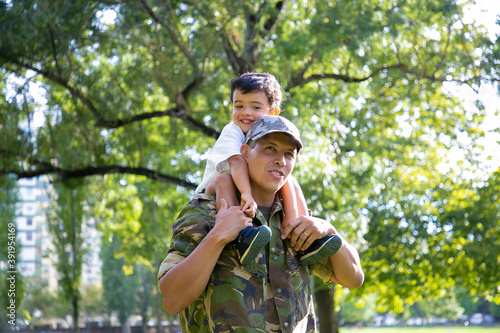 Loving father holding son on neck and walking in city park. Happy Caucasian son sitting on neck of dad in uniform, hugging him and looking away. Family reunion, fatherhood and returning home concept