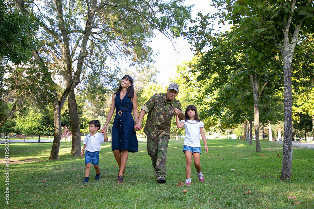Obraz premium Front view of happy family walking together on meadow in park. Father wearing military uniform and showing something to daughter. Long-haired mom smiling. Family reunion and returning home concept