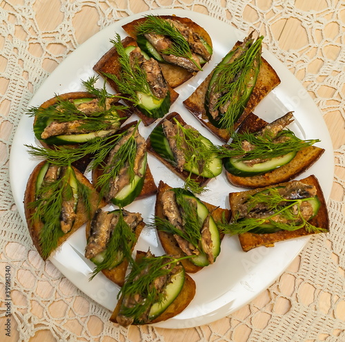 Sandwiches of toasted slices of rye bread, fresh cucumber, sprats from Baltic sprat and dill sprigs on a white plate closeup