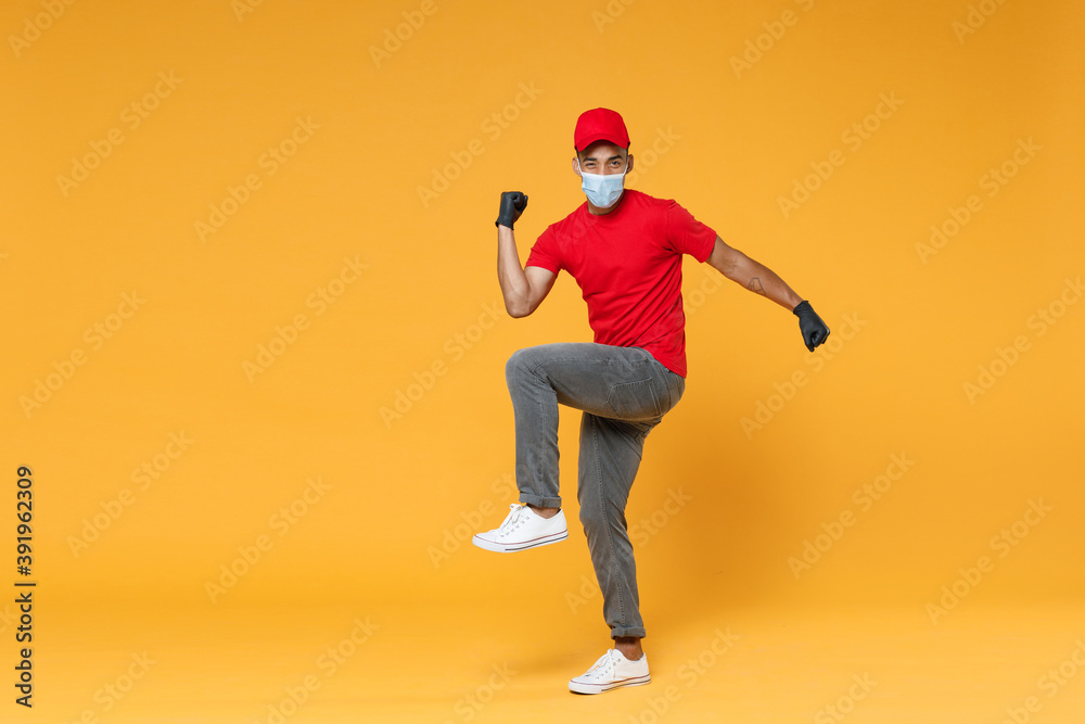 Full length delivery employee african man in red cap t-shirt face mask gloves uniform work courier dealer service on quarantine coronavirus covid-19 virus concept isolated on yellow background studio.