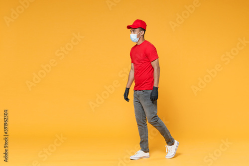 Full length delivery employee african man in red cap t-shirt face mask gloves uniform work courier dealer service on quarantine coronavirus covid-19 virus concept isolated on yellow background studio.